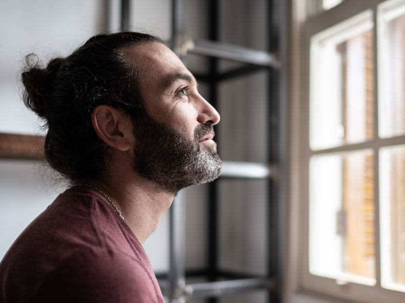 man with long hair and a beard smiling while looking out a window into sunlight