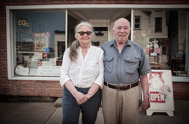 An elderly couple stand in an art studio surrounded by botanical paintings