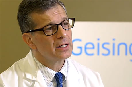A man wearing glasses and a white lab coat speaks, with a partially visible "Geisinger" sign in the background.