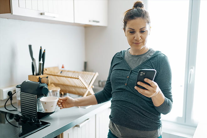 A pregnant woman prepares coffee in her kitchen as she looks at her cell phone.